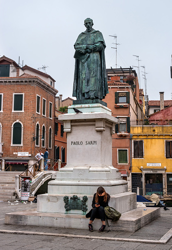 Venice, Italy - October 13, 2017: Statue of Paolo Sarpi in Venice. At the foot of the monument sits a girl. She has a phone in her hands. She looks at the phone screen.