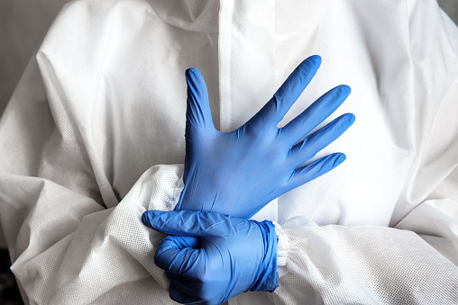 Doctor or nurse in white medical PPE suit puts on protective gloves, physician in hospital close-up. Concept of professional clinical care, uniform and healthcare during COVID-19 coronavirus pandemic