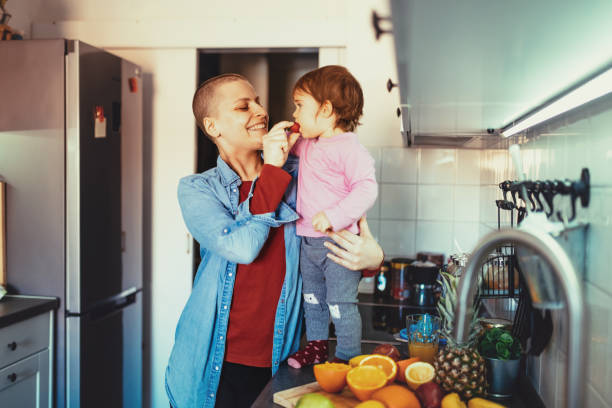 Cancer mother making breakfast with daughter Day in a life of cancer patient - portrait of woman at home and office after chemotherapy due to lymph nodes cancer. Woman is 30 year old real cancer cured patient, photographs and videos taken weeks after last chemotherapy session with visible burns on woman's neck and on facial skin. food chain stock pictures, royalty-free photos & images