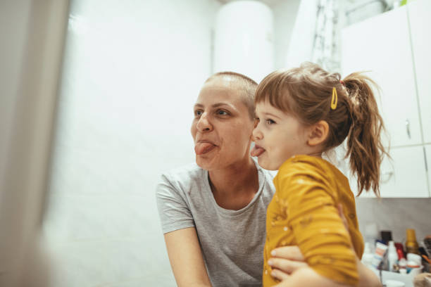 Mother with cancer hugging daughter in bathroom and making faces. Day in a life of cancer patient - portrait of woman at home and office after chemotherapy due to lymph nodes cancer. Woman is 30 year old real cancer cured patient, photographs and videos taken weeks after last chemotherapy session with visible burns on woman's neck and on facial skin. cancer illness stock pictures, royalty-free photos & images