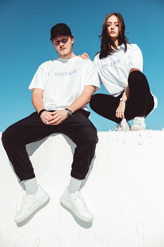 Young fashionable couple wearing designer t-shirts (property released) and black pants sitting outdoors on concrete wall, looking towards the camera. Shot against blue summer sky. Millennial Generation Male - Female Young Urban Fashion Portrait,