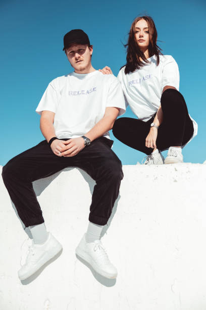 Fashionable young couple sitting on concrete wall Modern Fashion Portrait Young fashionable couple wearing designer t-shirts (property released) and black pants sitting outdoors on concrete wall, looking towards the camera. Shot against blue summer sky. Millennial Generation Male - Female Young Urban Fashion Portrait, street fashion stock pictures, royalty-free photos & images