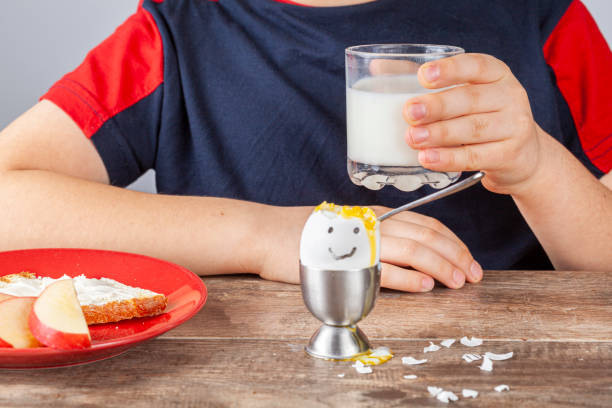 Breakfast table with simple balanced nutritious ingredients. A soft boiled egg with face painted on, a slice of toasted bread with cream cheese spread, apple, a glass of milk with a boy drinking it.