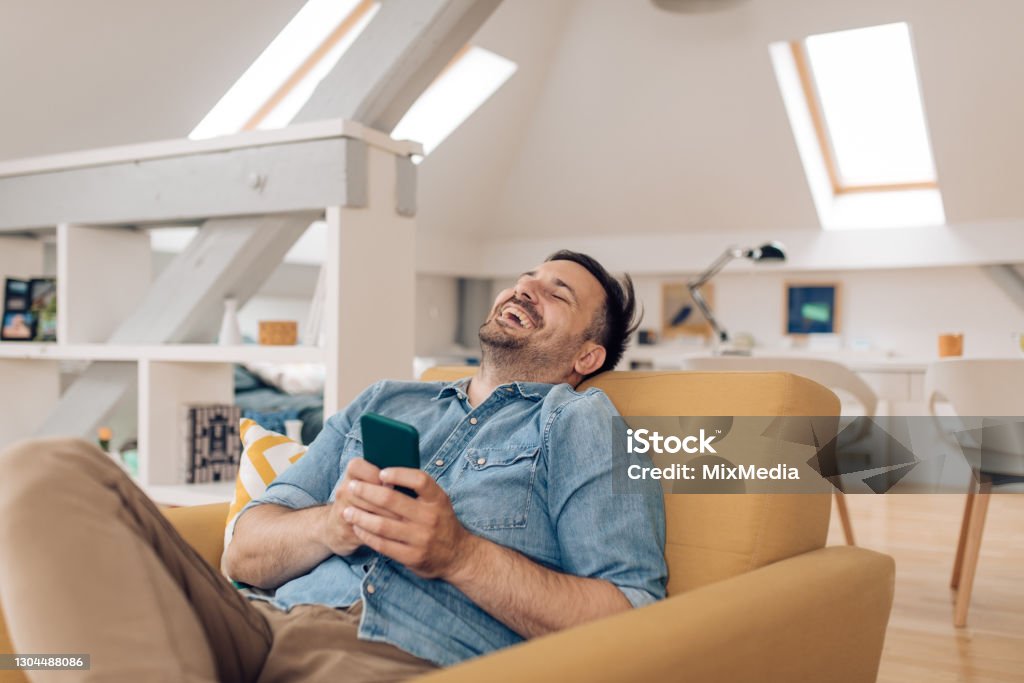 Portrait of a man laughing while watching somethi9ng online Young man relaxing and using a smartphone at home. 30-34 Years Stock Photo