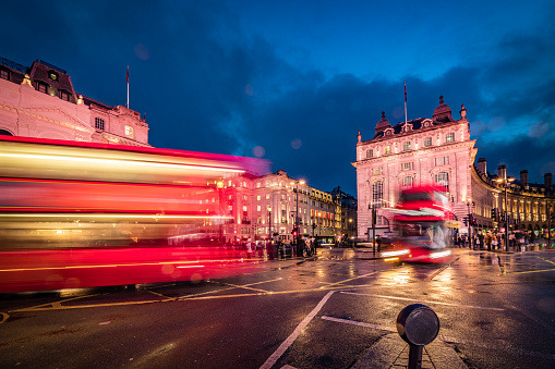 Long exposure photo with evening illumination and reflections from the wet roads at Piccadilly Circus in London city during blue hour on a rainy English evening. Crowd and traffic with motion blur due to the exposure time with unrecognisable accidental commuters and tourist. Image has copy space and it is ideal for background. Shot on Canon EOS R full frame system.