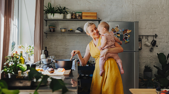 A beautiful young happy Caucasian mother holding her adorable baby girl in her arms while preparing lunch with a mixing spoon over a kitchen stove at home.