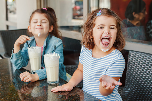 Two funny Caucasian little preschool sisters siblings drink milk shakes in cafe. Friends girls having fun together. Cold summer desserts for kids. Happy authentic childhood lifestyle.