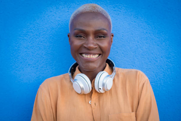 an adult black woman poses with headphones on a blue wall and smiles stock photo
