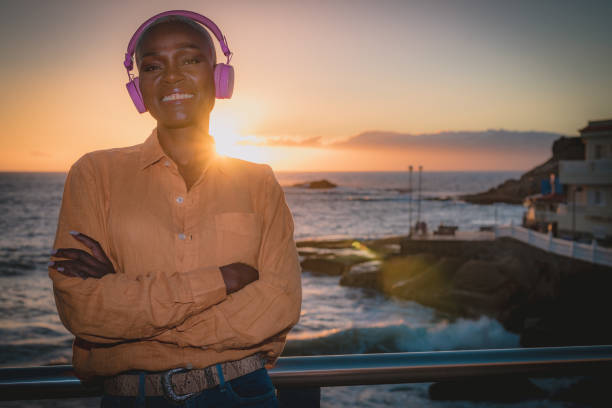 a black adult woman is listening to happy music, she sees the sunset and the sun rays with the sea in the background, she is relaxed and happy. Focus on the face stock photo