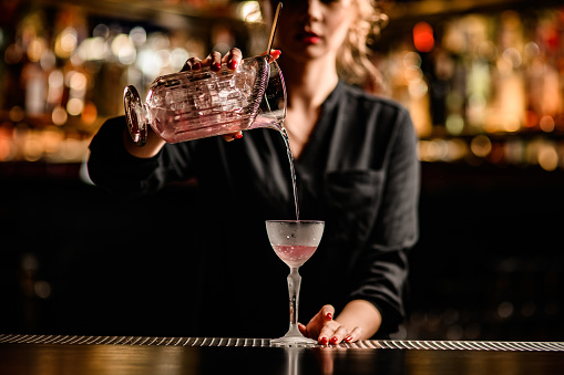 Close-up view of beautiful young female bartender pouring cocktail from mixing cup into crystal glass. Blurred background