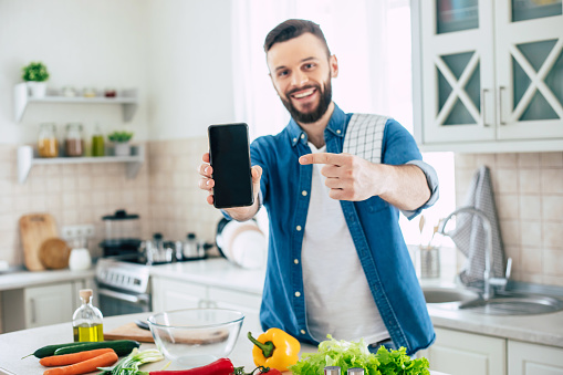Happy bearded man in the kitchen at home while he is preparing the healthy fresh vegan salad and pointing on the smart phone