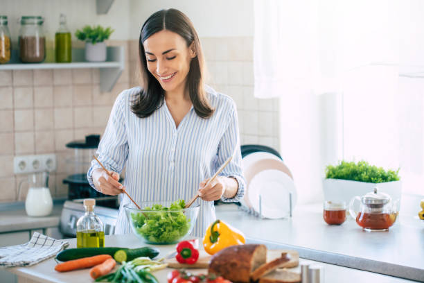 Happy smiling cute woman is preparing a fresh healthy vegan salad with many vegetables in the kitchen at home and trying a new recipe stock photo