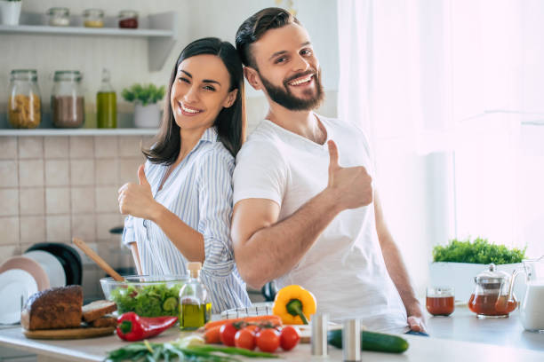 Excited smiling young couple in love making a super healthy vegan salad with many vegetables in the kitchen and showing thumbs up stock photo