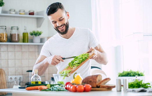 Handsome happy bearded man is preparing wonderful fresh vegan salad in the kitchen at home