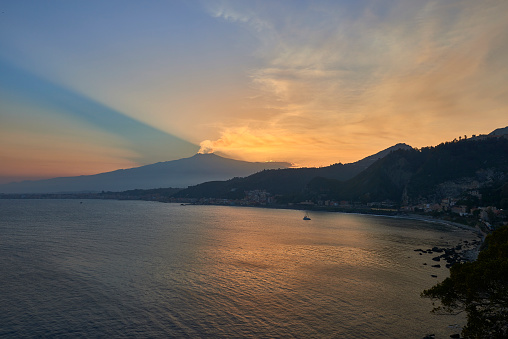 the bay of Taormina at sunset with the  smoking Etna Volcano dominating the scene while the sky is colored