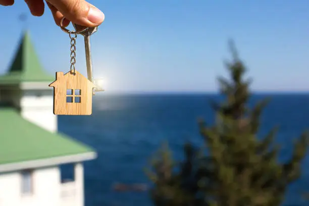 Photo of Wooden pendant of a house and key. Background of sea, fir tree and cottage. House and life on ocean, construction, project, moving to new house, mortgage, rent and purchase of real estate. Copy space