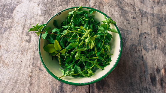 Fresh cut organic basil and mint leaves in a yellow enamel tin bowl on a wooden tabletop