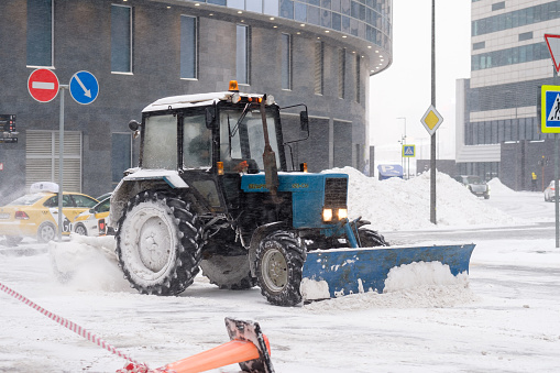 Moscow. Russia. February 12, 2021. A tractor with a bucket and a mechanical rotating brush sweeps snow on a street in a city during a snowfall. The technique is covered with a thick layer of snow.