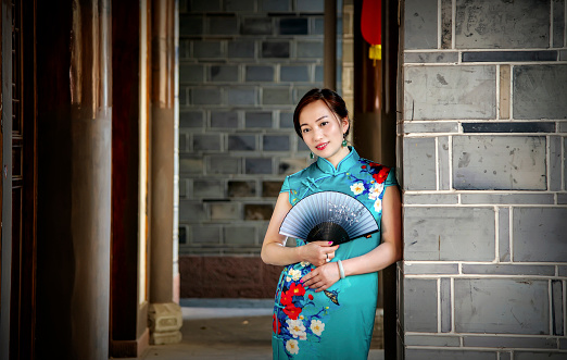 A woman dressed in a cheongsam stood in front of the gate of an ancient house, holding a fan in her hand.Retro color card style