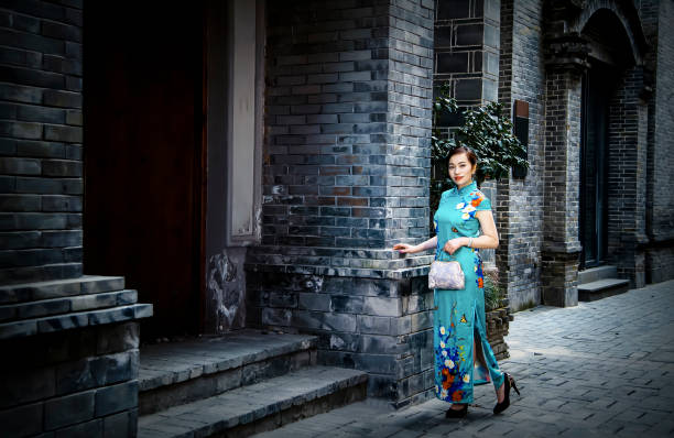 A woman in a cheongsam, holding a handbag, is standing in front of an ancient house.Retro color card style