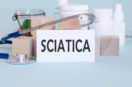 SCIATICA words written on white medical card, with stethoscope, green flower, medical pills and wooden blocks on blue background. Medical and healthcare concept. Selective focus