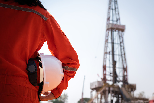 A worker is holding safety hardhat or helmet with blurred background of drilling rig derrick structure, selective focus at hardhat. Ready to working in oil field industrial concept photo.