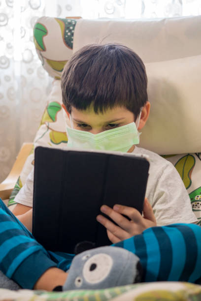 istock Small Child in a Mask To Avoid Covid-19 Coronavirus while Playing with Digital Tablet 1304545344