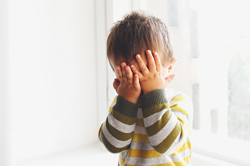 Portrait of adorable little boy sitting on the windowsill and crying. Upset child covering his face at home. Barefoot kid hiding behind palms of his hands. Close up, copy space, background.