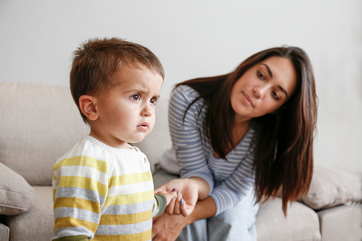 Portrait of adorable little boy sitting on the textile couch with his mom and crying. Upset toddler throwing a tantrum at home. Barefoot calling for attention. Close up, copy space, background.