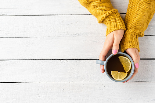 Woman’s hands in knitted sweater holding mug of tea with lemon on white wooden background with copyspace. Top view