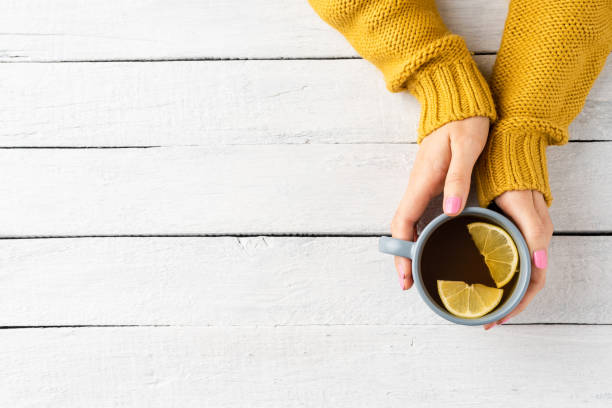Woman’s hands in knitted sweater holding mug of tea with lemon on white wooden background with copyspace. Top view stock photo
