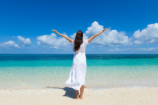 Woman in white dress posing in tropical sea beach with arms raised