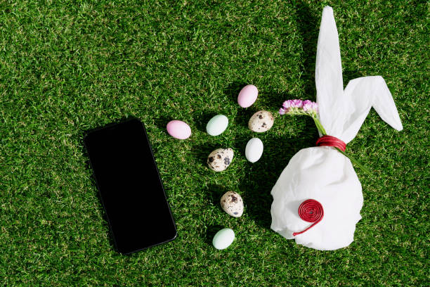 istock Chocolate Easter eggs glazed with pastel coloring and quail eggs over green grass background. Bunny shaped white baking paper. Black cell phone. Happy Easter concept. Copy space. Top view 1304558366