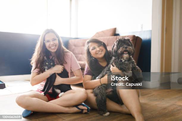 Portrait Of Two Smiling Young Women Sitting On Living Room Floor Holding A Cat And A Dog Stock Photo - Download Image Now