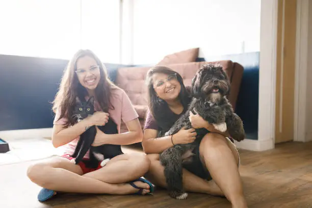 Photo of Portrait of two smiling young women sitting on living room floor holding a cat and a dog