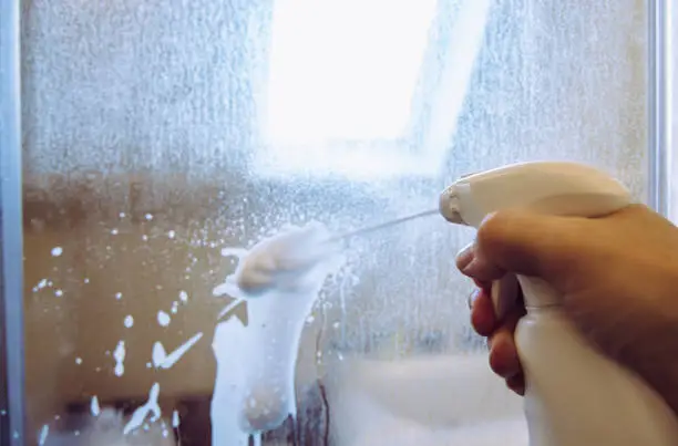 Photo of Person hand using limescale remover chemical foam. Descaling shower case glass door. Work in progress.