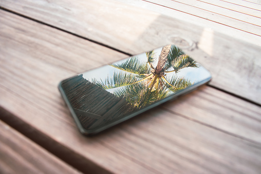 Teléfono móvil con reflejo de palmera en exhibición. Smartphone sobre mesa de madera en complejo tropical. Concepto de vacaciones. Reflexión de palmeras en la pantalla del teléfono. photo