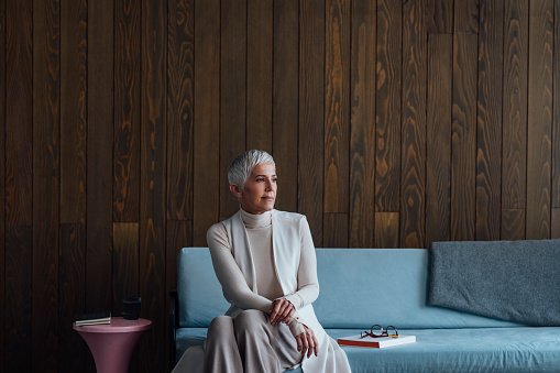 A portrait of a senior woman architect wearing a white turtleneck, sitting on a couch in her studio.