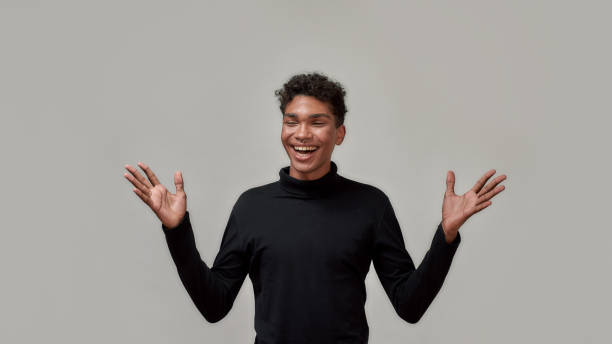 istock Happy young guy in black turtleneck laughing with eyes closed while standing with raised hands isolated over gray background 1304575314