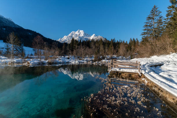 romantico sentiero attraverso la neve lungo un bellissimo lago vulcanico color smeraldo e limpido in inverno in una riserva naturale turistica delle alpi. riflesso di montagne innevate che brillano sotto il sole nascente. - european alps cold mountain range clear sky foto e immagini stock