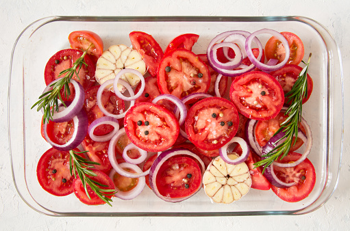 Top view of sliced tomatoes, garlic, onion, and rosemary in a glass oven dish. Mediterranean food