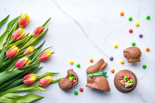 Top down view on delicious chocolate Easter bunny, hen and colorful candy next to bouquet of yellow red tulips. Easter concept, space for text in the middle.