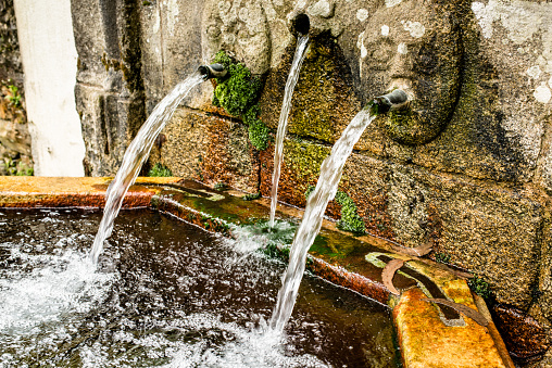 Stone fountain decorated with human faces pouring water into a sink.