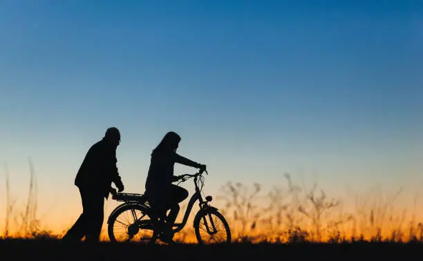 Silhouette of a girl and man on the e-bike or electric bicycle on the sunset background. Country style, transportation in the village. Copy space.Travel, father and daughter.