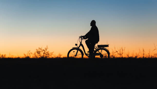 Male cyclist on the e-bike or electric bicycle on the sunset background riding up the hill. Silhouette of the old man in profile. Active pension. Travel. Sport. stock photo