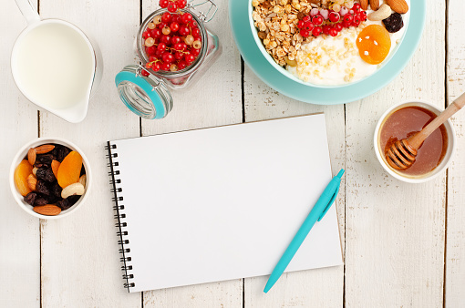 Notepad for weight loss plan, healthy breakfast of granola bowl on white wooden background. Top view