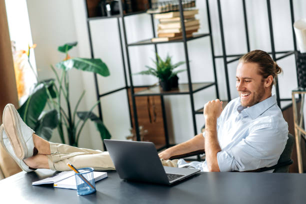 istock Smiling attractive young man, freelancer or manager, is relaxing at workplace with legs thrown on table. Modern guy uses laptop, browses the Internet, looks for ideas for a project, while sitting at home office 1304609928