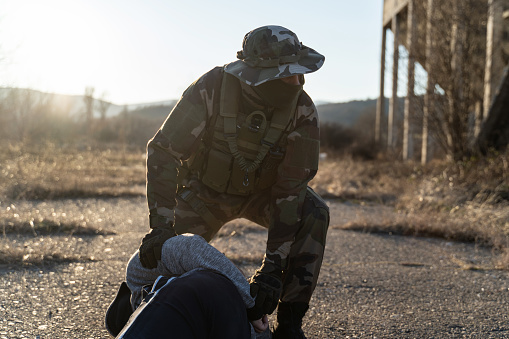 Soldier in military uniform arresting a terrorist or illegal immigrant during border patrol mission - special force holding a criminal or migrant on the ground in rural environment in sunny day
