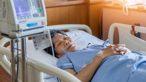 Hospitalized elderly patient senior woman sleeping on bed in hospital ward room with iv medical infusion pump infusing saline solution fluids, medication medicine for health treatment recovery