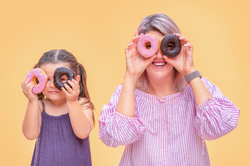 Funny young woman and child on yellow wall background.  Mother and her daughter girl have fun with colorful donuts.  Pink and chocolate donuts.  Junk food diet.